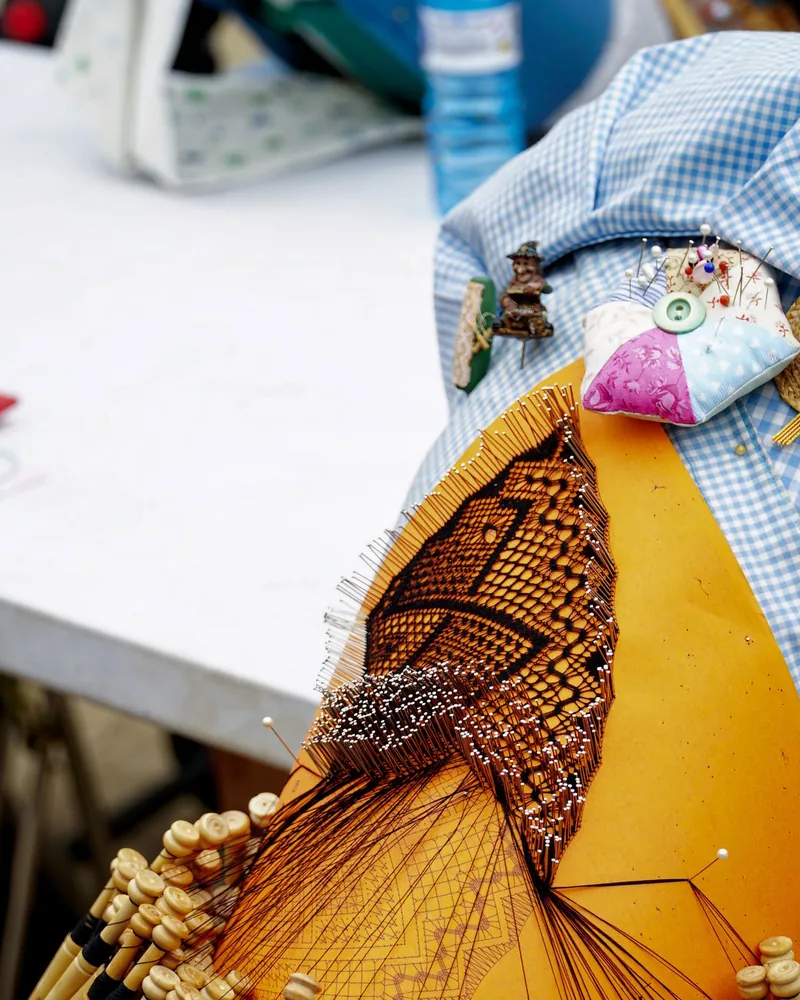 Detalle de una labor de encaje de bolillos en curso sobre un picado naranja, con alfileres clavados y bolillos colgando