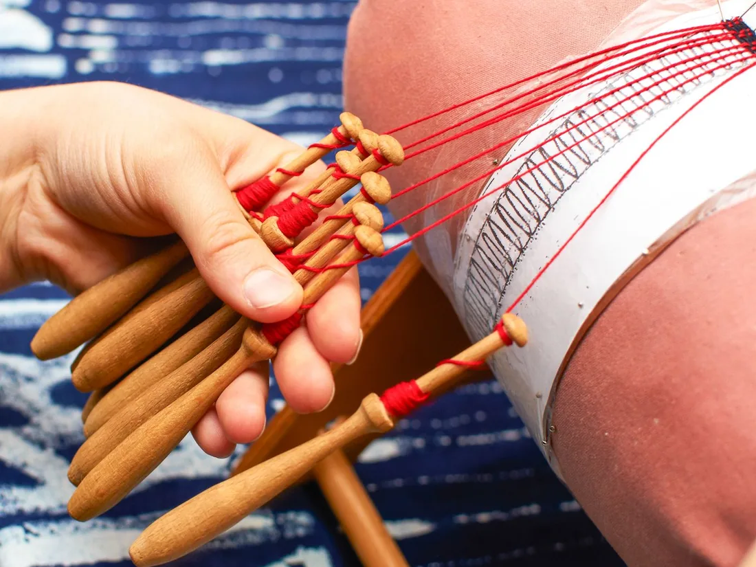 Manos sosteniendo un grupo de bolillos de madera con hilo rojo sobre un mundillo cubierto de alfileres